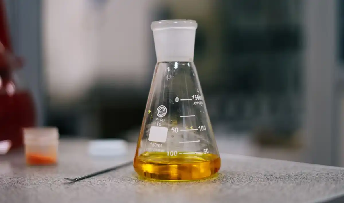 Um frasco Erlenmeyer de vidro contendo um líquido amarelo, colocado sobre uma bancada de laboratório ao lado de uma pipeta e pequenos recipientes. Esses frascos são comumente utilizados em química e microbiologia para misturar, aquecer e armazenar soluções durante experimentos.