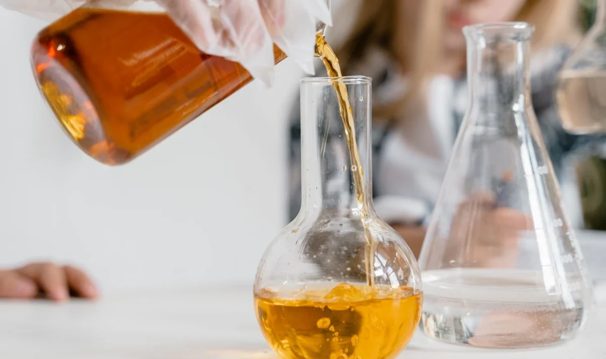 pessoa usando luvas transparentes enquanto põe um líquido amarelo em um frasco de vidro em um laboratório.