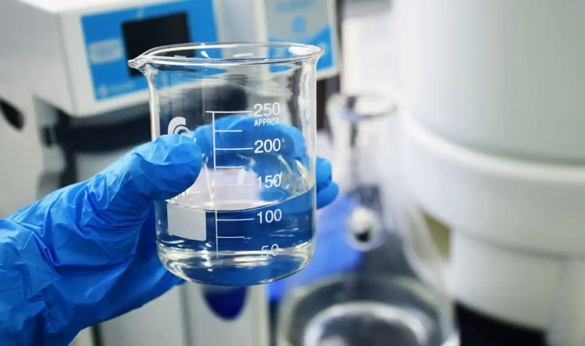 Uma mão enluvada segura um béquer de vidro transparente contendo um líquido transparente em um ambiente de laboratório. O fundo mostra equipamentos científicos, sugerindo um ambiente de pesquisa ou análise.