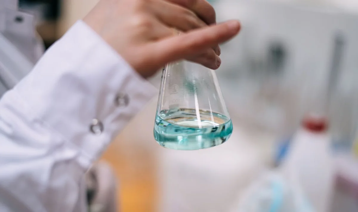 Cientista de jaleco branco segurando um frasco Erlenmeyer de vidro transparente contendo um líquido azul. A imagem destaca o uso de vidraria de laboratório em experimentos e pesquisas químicas.