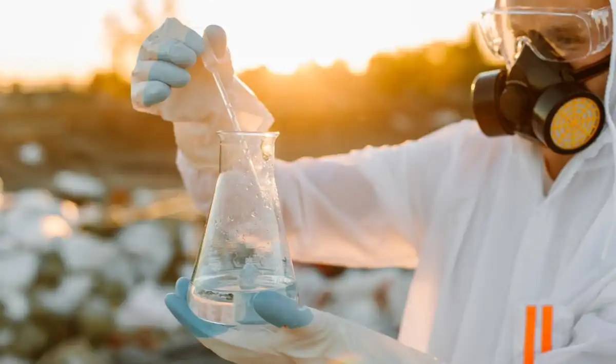 Cientista com equipamento de proteção individual manuseando um béquer de vidro contendo um líquido transparente durante um teste ambiental ao ar livre.