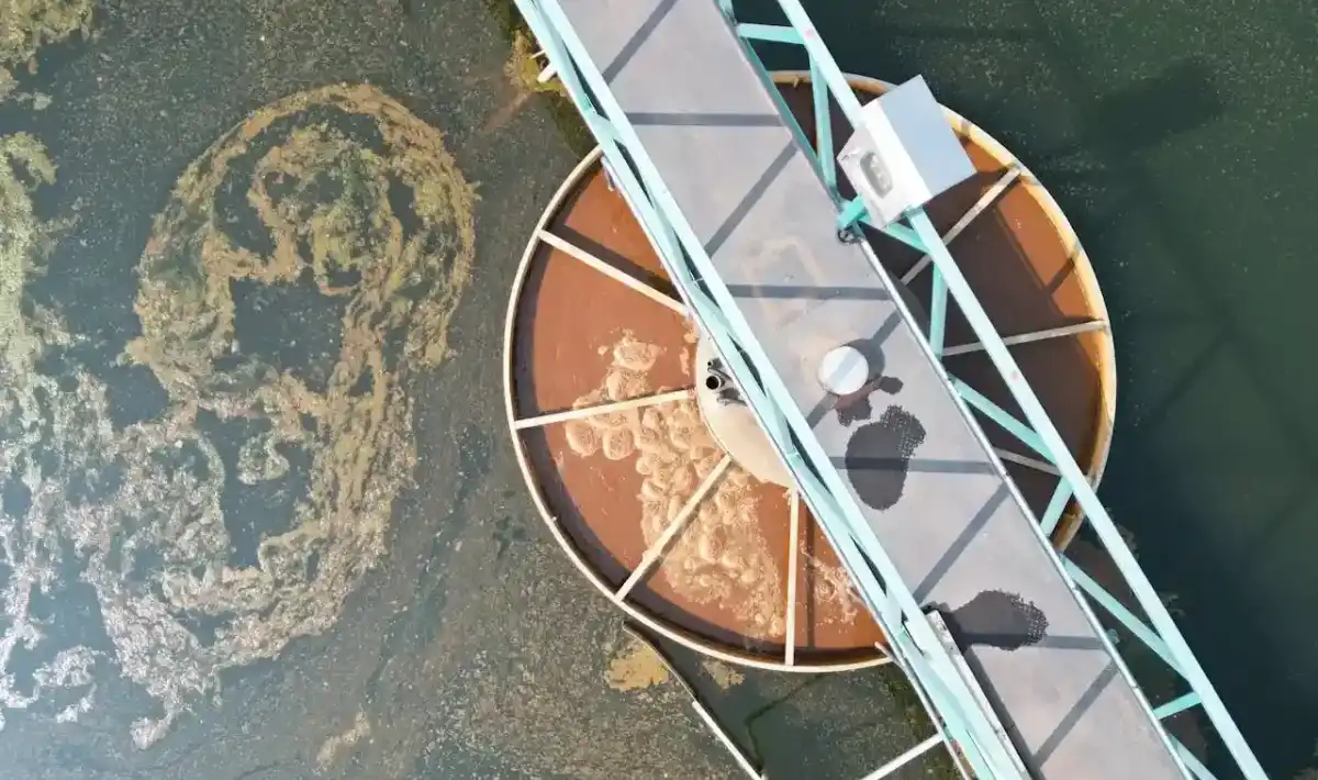 A fotografia aérea mostra uma estação de tratamento de águas residuais com um tanque de decantação circular e uma passarela para acesso de manutenção.