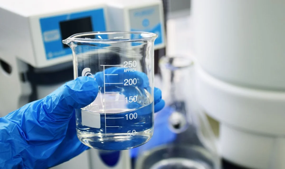 Uma mão enluvada segura um béquer de vidro contendo um líquido límpido em um ambiente de laboratório. Ao fundo, instrumentos científicos e vidrarias sugerem a realização de testes químicos ou analíticos.